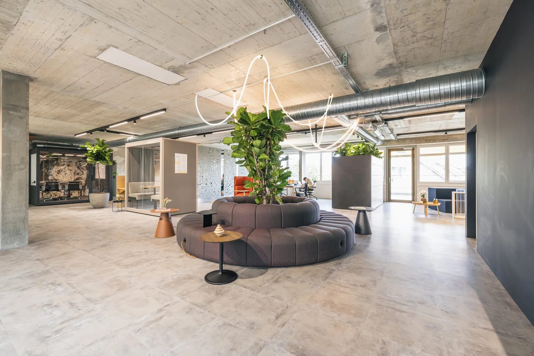 Offene Fläche der Work Factory mit rundem Sofa um einen Baum, Rückzugsraum im Hintergrund